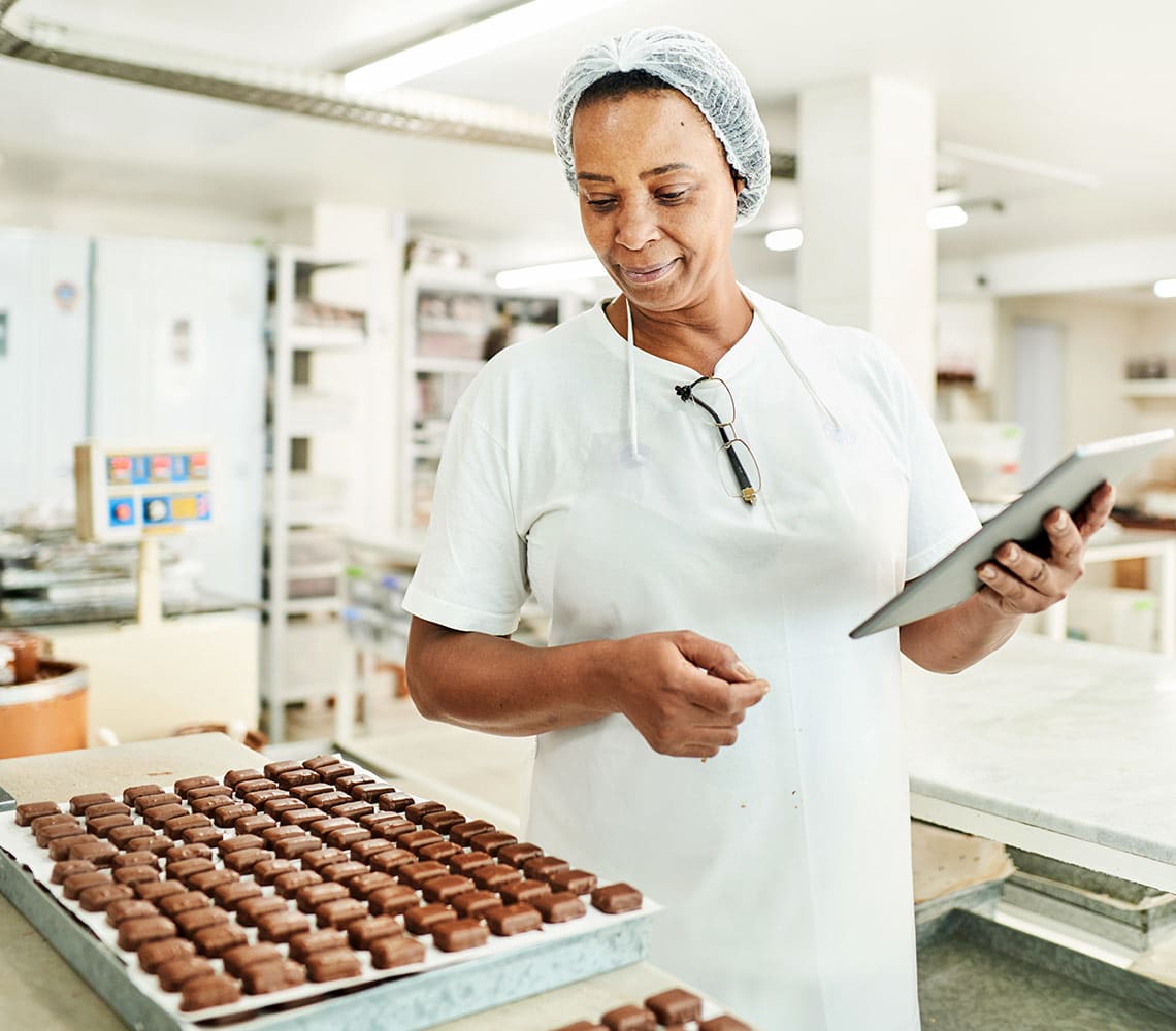 A woman in a white uniform and hair net is holding a tablet while examining the freshly made chocolates.